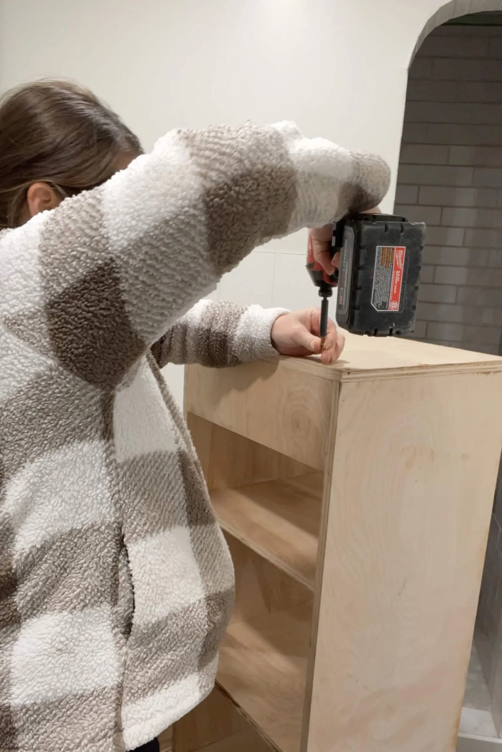 Woman in a cozy plaid fleece using a power drill to secure the top panel of a custom-built wooden storage unit. The cabinet is partially assembled, featuring multiple shelves, as she works in a bright, tiled space.